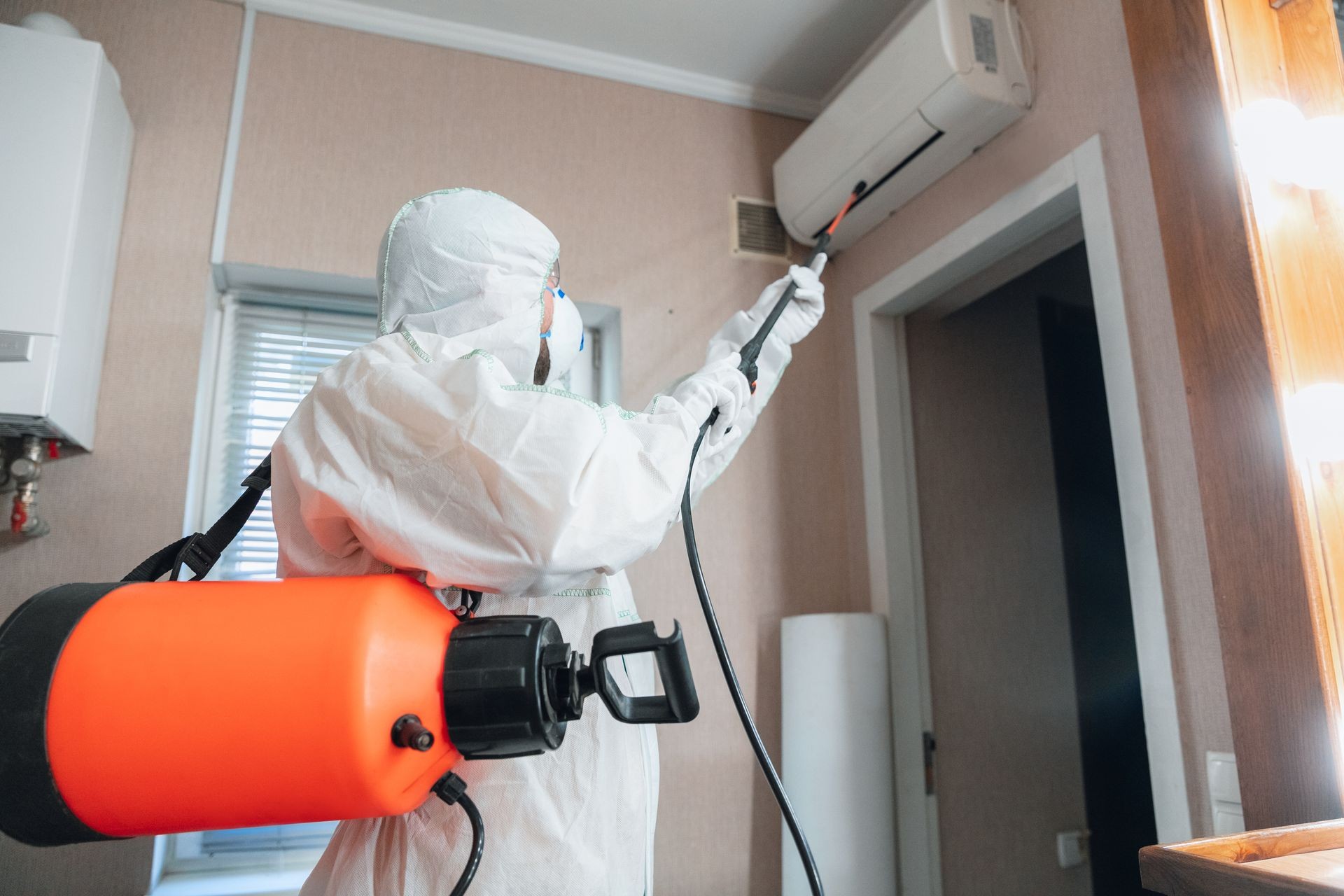 Person in protective suit spraying disinfectant on an air conditioning unit indoors.