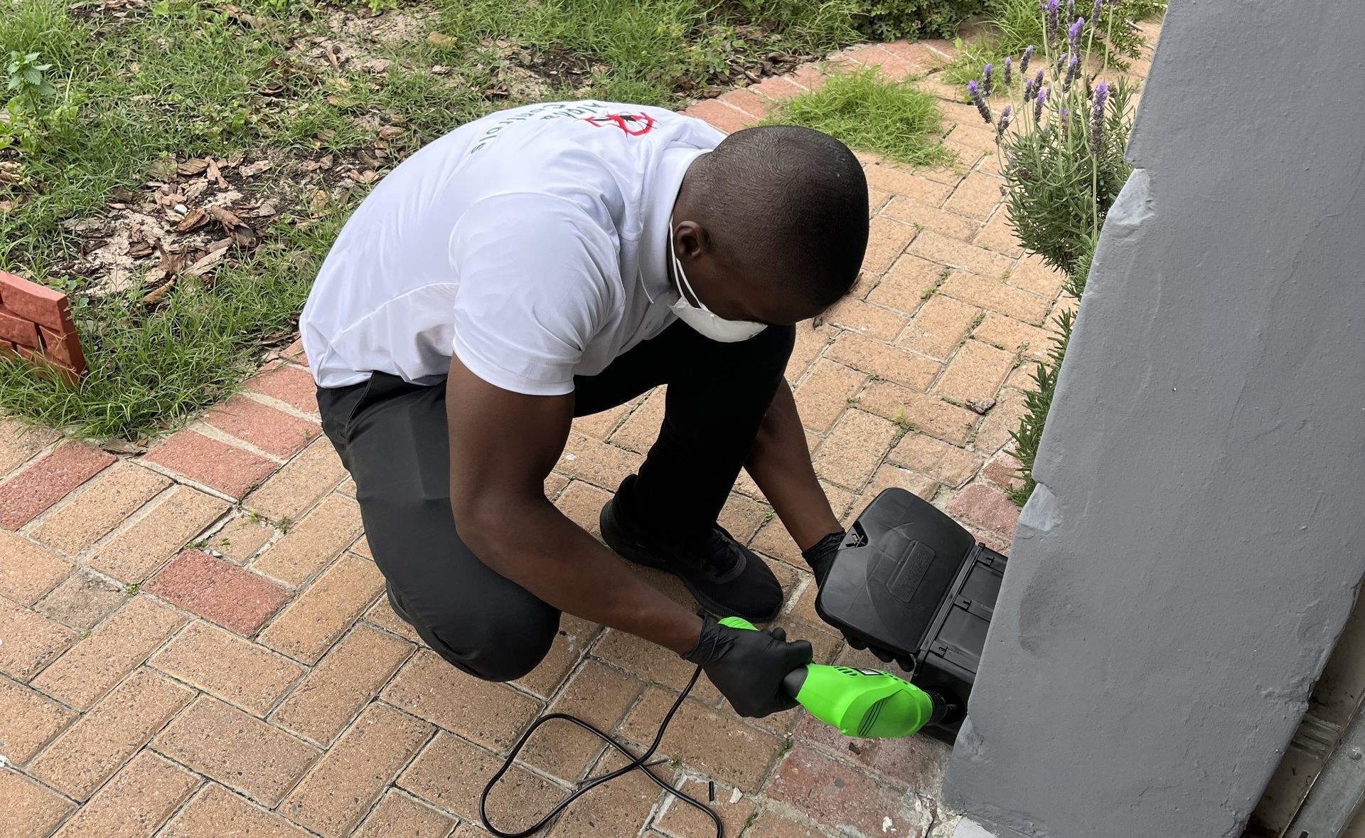 Person wearing gloves and a mask using a green hand tool on a brick pathway near a building.